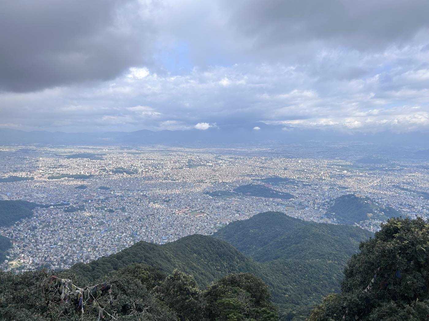 Panoramic view over the Kathmandu Valley and its sprawling cityscape from a forested hilltop, prayer flags in the foreground